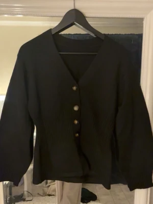 Svart ribbad kofta med knappar - Snygg svart kofta med v-ringning och fyra knappar framtill. Koftan har ribbade partier i midjan som ger en schysst passform och ballongärmar med ribbade muddar. Perfekt att slänga över en t-shirt eller topp för en chill look.