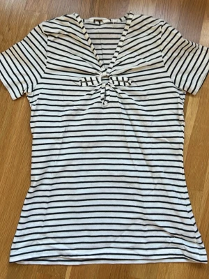 Randig topp med knut från NAF NAF - Snygg vit topp med svarta horisontella ränder från NAF NAF. Toppen har korta ärmar, v-ringning och en dekorativ knut framtill. Tillverkad i mjukt, tunt material som känns skönt mot huden. Perfekt för en avslappnad och trendig look.