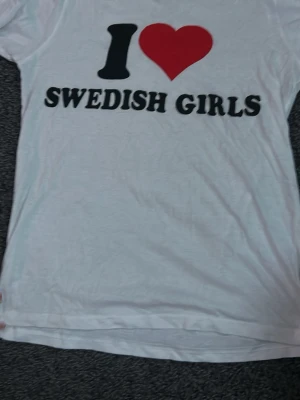 Vit t-shirt med tryck I ❤️ SWEDISH GIRLS - Vit t-shirt med svart och rött tryck där det står 'I ❤️ SWEDISH GIRLS' på framsidan. Klassisk passform och rund halsringning. Perfekt för dig som vill sticka ut med ett roligt statement-plagg.