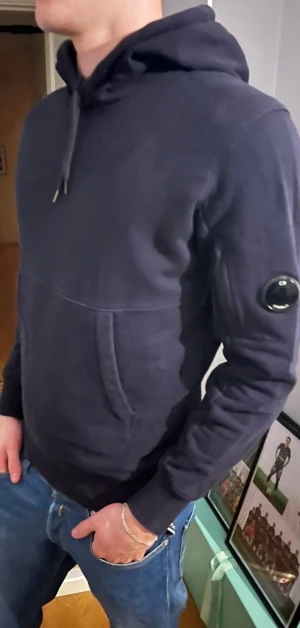 Mörkblå hoodie från C.P. Company - Säljer en mörkblå hoodie från C.P. Company med klassisk huva och dragsko. Ikonisk linsdetalj på vänster ärm och logga på huvan. Tillverkad i mjukt bomullsmaterial och har en stor magficka framtill. Perfekt för en avslappnad streetwear-look. Pris kan diskuteras, hör av er vid frågor! Nypris 3000kr. 