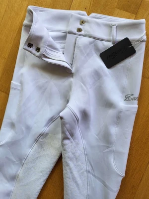 Vita ridbyxor från Zaczess - Snygga vita ridbyxor från Zaczess med helskoning i silikon för extra grepp. Byxorna har hög midja, dubbla knappar och diskreta fickor. Materialet är stretchigt och följsamt, perfekt för ridning och träning.