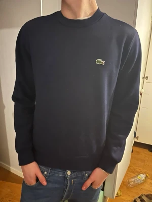 Mörkblå sweatshirt från Lacoste - Klassisk mörkblå sweatshirt från Lacoste med rund halsringning och den ikoniska krokodilloggan på bröstet. Tröjan har långa ärmar och ribbade muddar vid ärmslut och nederkant. Perfekt till jeans för en clean och stilren look. Pris kan diskuteras, hör av er vid frågor! Nypris 1200kr