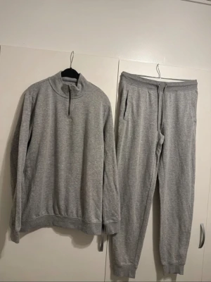 Grått loungewear-set med dragkedja - Grått loungewear-set med långärmad tröja och matchande byxor. Tröjan har halv dragkedja vid halsen och ribbade muddar. Byxorna har dragsko i midjan, fickor och ribbade benslut. Mjukt material, troligen bomull eller bomullsblandning.