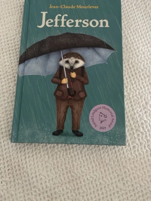Jefferson av Jean-Claude Mourlevat - En illustrerad bok med hård pärm, turkos omslag och motiv av en igelkott i brunt kostym som håller ett paraply i regnet. Boken är skriven av Jean-Claude Mourlevat och har en ALMA-pris-sticker på framsidan.