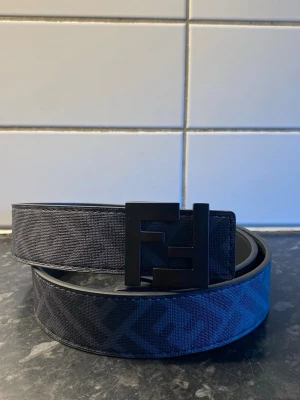 Svart bälte med FF-logga - Snyggt svart bälte med stor FF-logga i matt svart metall som spänne. Bältet har ett diskret mönster med FF-loggor längs hela bandet och är gjort i skinn med textil på ovansidan. Perfekt för att lyfta vilken outfit som helst.