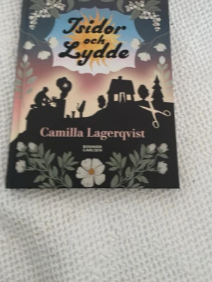 Isidor och Lydde av Lagerqvist - Romanen 'Isidor och Lydde' av Camilla Lagerqvist har ett färgstarkt omslag med siluetter, blommor och en dramatisk solnedgång. Boken är inbunden och riktar sig till unga vuxna. Perfekt för dig som gillar äventyr och vänskap.