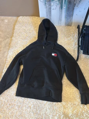 Svart hoodie från Tommy Jeans - Säljer en svart oversized hoodie från Tommy Jeans med klassisk logga på bröstet. Hoodien har en stor ficka framtill och justerbar dragsko i huvan. Materialet är mjukt och skönt, perfekt för en avslappnad stil. Passar dig som gillar streetwear och enkel design.