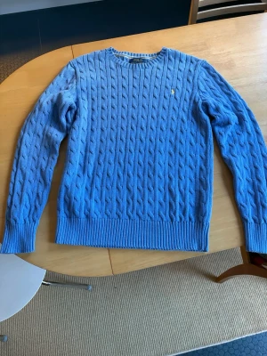 Blå stickad tröja Polo Ralph Lauren - Snygg blå kabelstickad tröja från Polo Ralph Lauren med rund halsringning och gul broderad logga på bröstet. Tröjan har ribbade muddar vid ärmslut och nederkant. Perfekt för dig som gillar klassisk preppy stil. Helt ny tröja