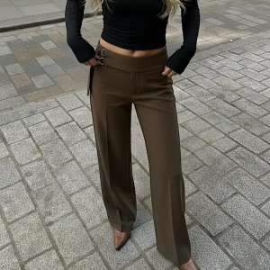 Bruna vida kostymbyxor med spänne - Säljer ett par bruna kostymbyxor med låg midja och vida ben. Byxorna har ett snyggt spänne i midjan på sidan som detalj. Perfekta för en trendig och stilren look.