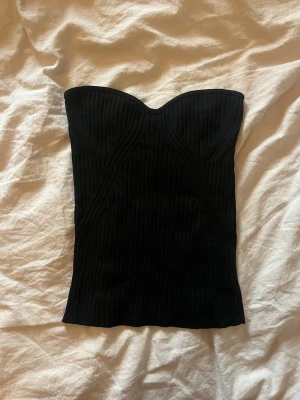 Svart ribbad tubtopp från Lindex - Snygg svart ribbad tubtopp från Lindex i storlek XS. Toppen är axelbandslös och har en croppad passform med markerad byst och stretchigt material som sitter bekvämt. Perfekt att styla med jeans eller kjol för en trendig look.