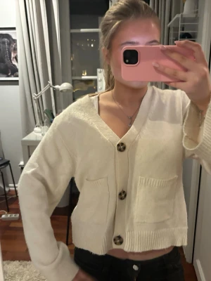 Krämvit kofta med stora knappar - Mysig krämvit kofta med v-ringning och tre stora, mönstrade knappar framtill. Koftan har två framfickor och ribbade muddar vid ärmslut och nederkant. Kortare modell som passar perfekt till jeans eller kjol.