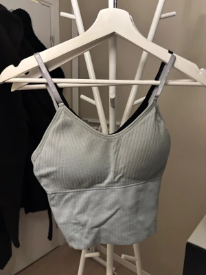 Ljusgrå ribbad body med smala band - Snygg ljusgrå body med ribbad struktur och smala axelband. Modellen har en enkel, stilren design med v-ringad halsringning och stretchigt material som sitter bekvämt mot kroppen. Perfekt att styla under skjortor eller tröjor.