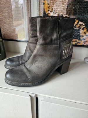 Svarta stövletter med nitar - Snygga svarta stövletter i läder med chunky klack och coola silverfärgade nitar på hälen. Modellen har rund tå och dragkedja på insidan. Perfekta för dig som gillar en edgy stil och vill ha boots som sticker ut.