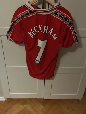 Manchester United Beckham tröja äkta! - Klassisk röd Manchester United matchtröja från Umbro med Beckham och nummer 7 på ryggen. Tröjan har vita och svarta detaljer, klubbmärke på bröstet och sponsorlogga från Sharp. Perfekt för dig som älskar fotboll och retrostil.