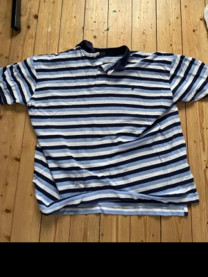 Randig pikétröja från Ralph Lauren - Snygg pikétröja från Ralph Lauren med klassiska blå och vita ränder. Tröjan har krage, korta ärmar och är tillverkad i 100% bomull. Perfekt för en avslappnad och stilren look.