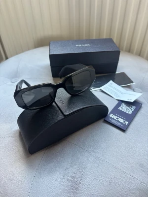 Svarta solglasögon från Prada - Snygga svarta solglasögon från Prada med rektangulära bågar och breda skalmar. Glasögonen har en modern och edgy look med diskret logga på sidan. Kommer med originalfodral och box, perfekt för dig som vill ha en stilren accessoar. Inte använda! Köptes på selfridges i Birmingham, UK.