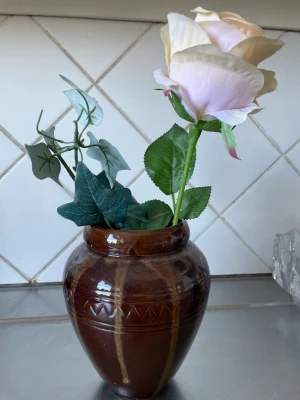 Brun keramikvas med konstblommor - Säljer en brun keramikvas med dekorativa mönster och konstblommor i form av en ljusrosa ros och gröna blad. Perfekt som inredningsdetalj för att ge rummet en fräsch känsla utan att behöva vattna blommor.
