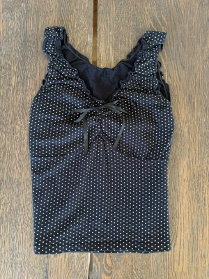 Svart prickig linnetopp med rosett - Snygg svart linnetopp med vita prickar och söt rosettdetalj framtill. Toppen har volangkant vid ringningen och är ärmlös med smala axelband. Perfekt för dig som gillar en feminin och lekfull stil.