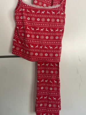 Röd julpyjamas med snöflingor & renar - Mysig röd pyjamas med vitt julmönster av snöflingor och renar. Setet består av ett linne med spetskant och långa byxor. Perfekt för vinterkvällar och julmys. Materialet känns mjukt och stretchigt, troligen bomullsblandning.