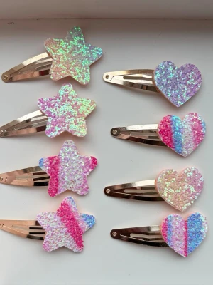 Glittriga hårspännen stjärna & hjärta - Säljer ett set med åtta glittriga hårspännen i form av stjärnor och hjärtan. Färgerna är rosa, blå, vit och regnbågsfärgade nyanser. Spännena har metallicbas och dekorationerna är täckta av skimrande paljetter. Perfekta för att piffa upp frisyren!