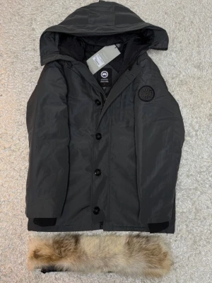 Svart parkajacka från Canada Goose - Parkajacka i svart från Canada Goose med avtagbar huva och pälsdetalj. Jackan har stora knappar framtill, patch med logotyp på bröstet och är tillverkad i slitstarkt material. Perfekt för kalla dagar och har en klassisk, rak passform.