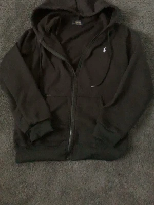 Svart hoodie från Polo Ralph Lauren - Svart hoodie med dragkedja från Polo Ralph Lauren. Klassisk design med vit broderad logga på bröstet, huva med dragsko och två fickor framtill. Tillverkad i mjukt bomullsmaterial som känns skönt mot huden. Perfekt för en avslappnad och stilren look.