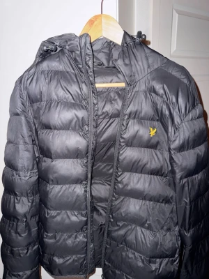 Svart pufferjacka från Lyle & Scott - Snygg svart pufferjacka från Lyle & Scott med huva och gul logga på bröstet. Jackan har quiltad design och dragkedja framtill. Perfekt för kalla dagar och riktigt skön att bära. Materialet är lätt och ger bra värme. Ett litet hål vid armhålan på högerarmen.