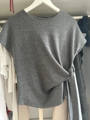 Grå stickad topp med knytband - Säljer en stilren grå stickad topp med rund hals och korta ärmar. Toppen har ett snyggt knytband i sidan som ger en cool detalj och markerar midjan. Perfekt att matcha med jeans eller kjol för en trendig look.