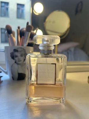 Coco Mademoiselle parfym från Chanel - Elegant parfymflaska från Chanel, modellen Coco Mademoiselle. Flaskan är i genomskinligt glas med en ljusrosa vätska och har en stilren, fyrkantig form med en frostad, geometrisk kork. Märkets logga och namn syns tydligt på etiketten framtill.