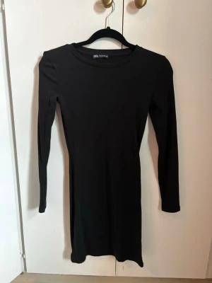 Svart långärmad klänning från Zara - En enkel och stilren svart klänning från Zara med lång ärm och rund halsringning. Klänningen är kort i modellen och har en tight passform som framhäver figuren. Perfekt basplagg att styla på olika sätt. Använd en gång.