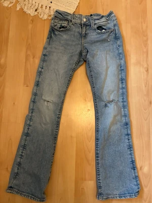 Ljusblå bootcut jeans med slitningar - Säljer ett par ljusblå bootcut jeans med slitningar på knäna. Jeansen har knapp- och dragkedjestängning samt ficklock med knappar där bak. Mycket bra skick. Storlek 164.