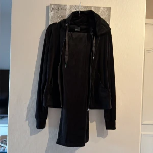 Svart mjukisset från Gina Tricot - Säljer en svart huvtröja i mjuk sammet från Gina Tricot. Tröjan har dragkedja framtill, stora fickor och en rejäl huva med breda snören. Byxorna är också i sammet och raka i modellen. Perfekt för en chill och avslappnad stil. 