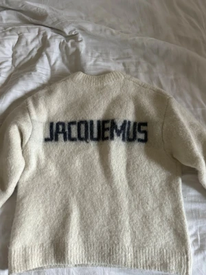 Vit stickad tröja från Jacquemus - Stickad tröja i mjuk ullblandning från Jacquemus. Tröjan är vit med rund halsringning och har ett stort Jacquemus-tryck i svart tryck på ryggen. Långärmad modell med ribbade muddar vid ärmslut och nederkant. Storleken är XL men passar M. Finns inga defekter på den.