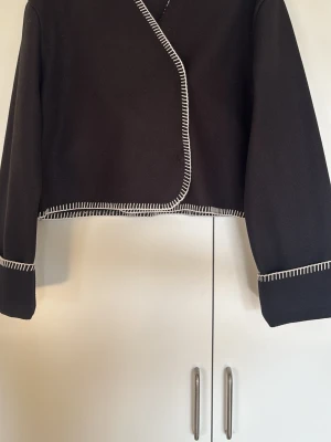 Svart jacka från Gina Tricot - Kort svart vårjacka från Gina Tricot med vita dekorativa sömmar längs kanterna. Koftan har lång ärm och öppen front med en knapp. Perfekt inför sommaren och våren