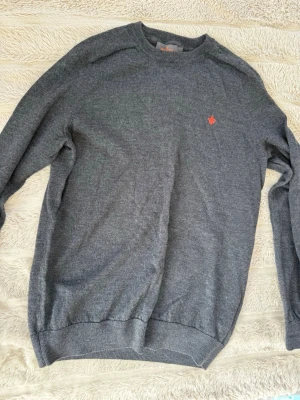 Grå stickad tröja från Morris - Säljer en stilren grå stickad tröja från Morris i 100% merinoull. Tröjan har rund halsringning, ribbade muddar och en liten orange logga broderad på bröstet. Perfekt för dig som gillar klassisk och enkel stil.