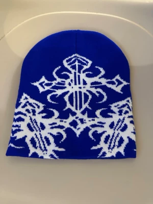 Blå mössa med vit unik design - Snygg blå mössa med vit unik design. Mössan har en klassisk form och är tillverkad i mjukt, stretchigt material som sitter skönt på huvudet. Perfekt för dig som vill sticka ut med en unik accessoar.