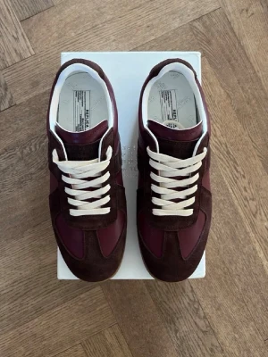  Maison Margielas Vinröda - Snygga sneakers i vinrött och mörkbrunt från Maison Margiela. Skorna har ovandel i mocka och läder, vita snören och klassisk gummisula. Perfekt för dig som gillar stilrena detaljer och en retrovibe.