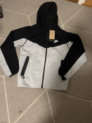 Nike grå och svart hoodie - Nike hoodie i grått och svart med dragkedja framtill och vit Nike-logga på bröstet. Hoodien har huva, långa ärmar och en ficka med dragkedja på sidan. Tillverkad i mjukt bomullsmaterial för en skön och avslappnad look.