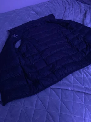 Svart dunväst från EA7 Emporio Armani - Snygg svart dunväst från EA7 Emporio Armani med klassisk quiltad design och diskret logga på bröstet. Västen har en stilren look och är lätt vadderad för extra värme. Perfekt att slänga över en hoodie eller tröja för en trendig streetwear-stil.
