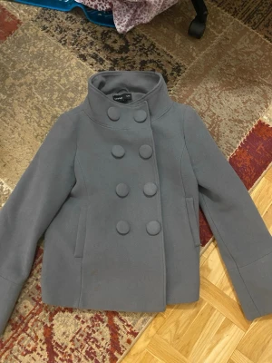 Grå dubbelknäppt kappa Sara Kelly - Stilren grå kappa från Sara Kelly by Ellos i storlek 36. Kappan har hög krage, dubbelknäppning med stora tygklädda knappar och två sidofickor. Materialet är ullblandning som ger en mjuk och varm känsla. Perfekt för kyliga dagar.