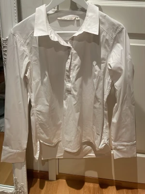Vit skjorta från strl M - Klassisk vit skjorta från House of Lola i storlek M. Skjortan har lång ärm, krage och knappar framtill. Perfekt basplagg med stilren look och normal passform.