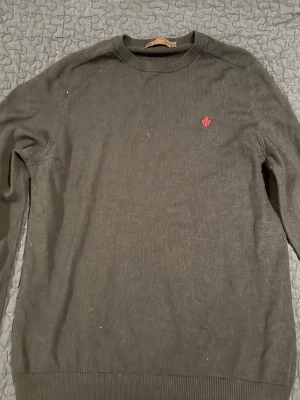 blå stickad tröja från Morris - Klassisk blå stickad tröja från Morris med rund halsringning och diskret röd logga broderad på bröstet. Tröjan har långa ärmar och ribbade muddar vid ärmslut och nederkant. Perfekt för en stilren och enkel look.