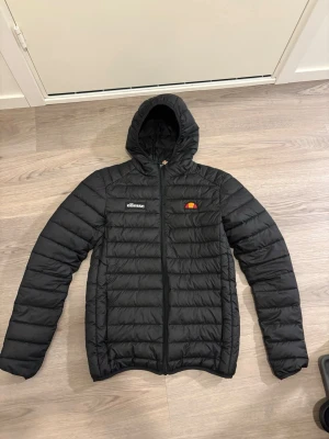 Svart pufferjacka från Ellesse - Snygg svart pufferjacka från Ellesse med huva och dragkedja framtill. Jackan har Ellesse-logga på bröstet och är quiltad för extra värme. Perfekt nu till våren och har en modern, sportig look.