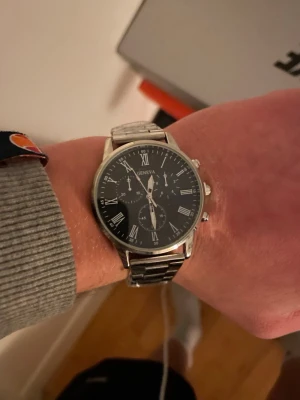 Klassisk silverfärgad Geneva klocka - Stilren klocka från Geneva med svart urtavla och romerska siffror. Armbandet är i blankt silverfärgat metall och har en klassisk länkdesign. Urtavlan har flera små visare och detaljer som ger en elegant look. Perfekt accessoar för dig som gillar tidlös stil.