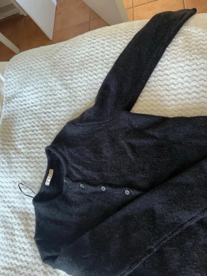 Svart kofta från Gina Tricot - Mysig svart kofta från Gina Tricot med rund halsringning och knappar framtill. Tillverkad i ett fluffigt, mjukt material som ger en avslappnad look. Perfekt att slänga över en topp eller t-shirt för extra värme.