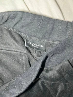  Juicy Couture XS - Säljer ett par svarta mjukisbyxor från Juicy Couture i velour. Byxorna har raka ben, bred resår i midjan och två fickor bak med knappdetaljer. Perfekta för en chill och trendig look.