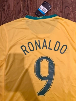 Brasilien Ronaldo 9 matchtröja Nike - Gul Brasilien matchtröja från Nike med gröna detaljer, nummer 9 och namnet Ronaldo på ryggen. Tillverkad i lätt och ventilerande material med Dri-FIT teknologi. Perfekt för fotbollsträning eller att bära som supporter.