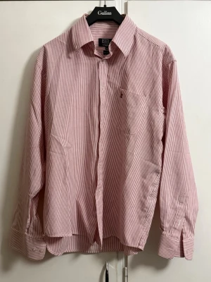 Rödvit randig skjorta från Polo Ralph Lauren - Klassisk skjorta från Polo Ralph Lauren i rödvitt randigt mönster. Skjortan har lång ärm, knappar framtill och en liten broderad logga på bröstet. Tillverkad i mjuk bomull och har en normal passform med klassisk krage.