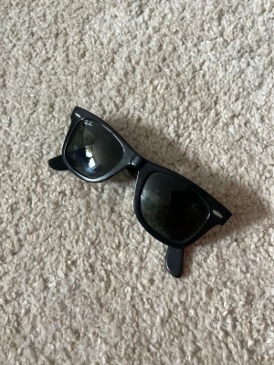 Svarta Ray-Ban Wayfarer solglasögon - Klassiska svarta Wayfarer solglasögon från Ray-Ban med ikonisk fyrkantig form och mörka glas. Snygg och tidlös design som passar till alla outfits. Ray-Ban-loggan syns tydligt på ena glaset. Ramen är i plast och glasen är mörkt tonade. Passar perfekt om man vill ha en lite lyxigare look till sommaren😊💕