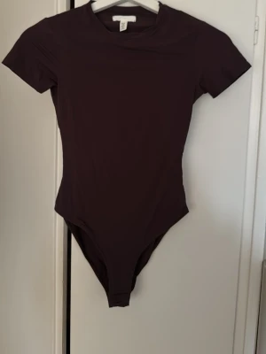 Mörkbrun body från H&M, XS - En enkel och stilren mörkbrun body från H&M i storlek XS. Bodyn har korta ärmar, rund halsringning och är tillverkad i mjukt stretchigt material som sitter snyggt mot kroppen. Perfekt att matcha med jeans eller kjol för en clean look. Använd en gång och mycket mörkare i verkligheten än på bild. 